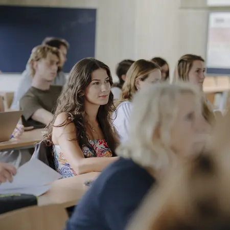 Eine Gruppe von Studierenden sitzt in einem Klassenzimmer und hört aufmerksam zu. Einige lächeln, während andere konzentriert auf den Vortragenden schauen. Die Atmosphäre ist freundlich und engagiert, mit einer Mischung aus verschiedenen Geschlechtern und Frisuren.