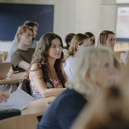 Eine Gruppe von Studierenden sitzt in einem Klassenzimmer und hört aufmerksam zu. Einige lächeln, während andere konzentriert auf den Vortragenden schauen. Die Atmosphäre ist freundlich und engagiert, mit einer Mischung aus verschiedenen Geschlechtern und Frisuren.
