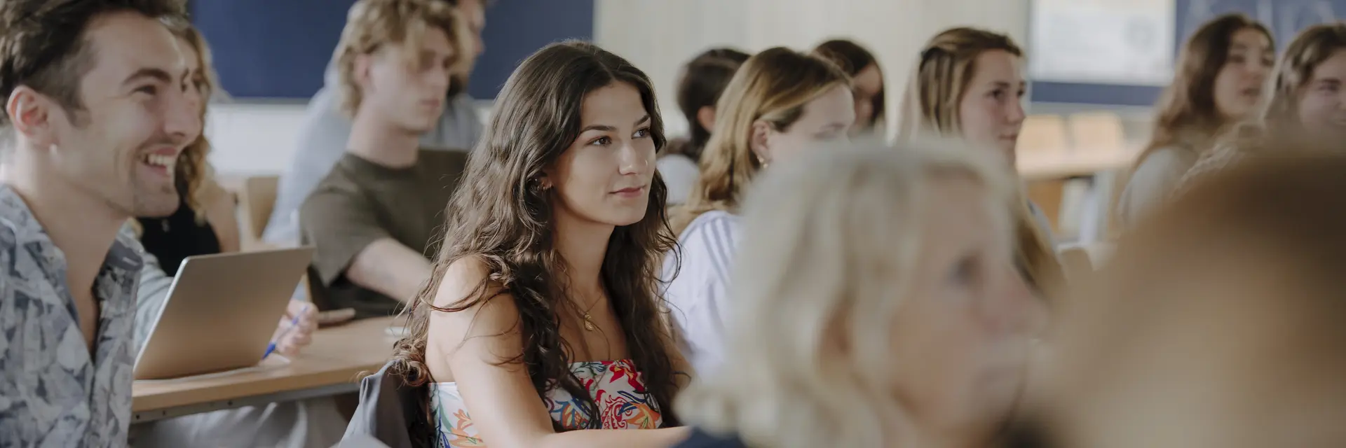 Eine Gruppe von Studierenden sitzt in einem Klassenzimmer und hört aufmerksam zu. Einige lächeln, während andere konzentriert auf den Vortragenden schauen. Die Atmosphäre ist freundlich und engagiert, mit einer Mischung aus verschiedenen Geschlechtern und Frisuren.