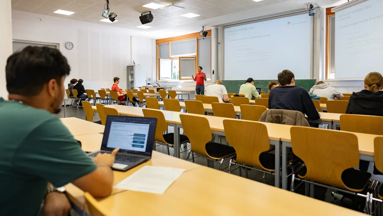 Ein Hörsaal mit mehreren Studierenden, die an ihren Tischen sitzen. Einige nutzen Laptops. Auf einer Leinwand vorne präsentiert eine Person Informationen. Der Raum ist hell und hat Fenster, durch die Tageslicht fällt.
