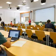 Ein Hörsaal mit mehreren Studierenden, die an ihren Tischen sitzen. Einige nutzen Laptops. Auf einer Leinwand vorne präsentiert eine Person Informationen. Der Raum ist hell und hat Fenster, durch die Tageslicht fällt.