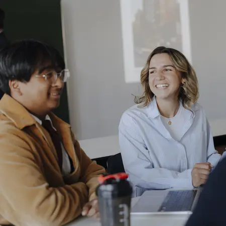 Eine Gruppe von jungen Menschen sitzt an einem Tisch und unterhält sich. Eine Frau mit blonden Haaren und einem hellen Hemd lächelt, während ein Mann mit Brille und einem braunen Jackett sie anblickt. Im Hintergrund stehen weitere Personen.