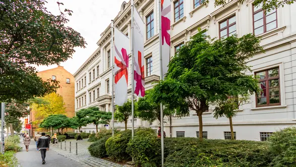 Eine Straße gesäumt von Bäumen und Sträuchern führt zu einem historischen Gebäude. Vor dem Gebäude stehen mehrere Fahnen mit bunten Mustern. Menschen gehen auf dem Gehweg, umgeben von einer ansprechenden Gartenlandschaft.