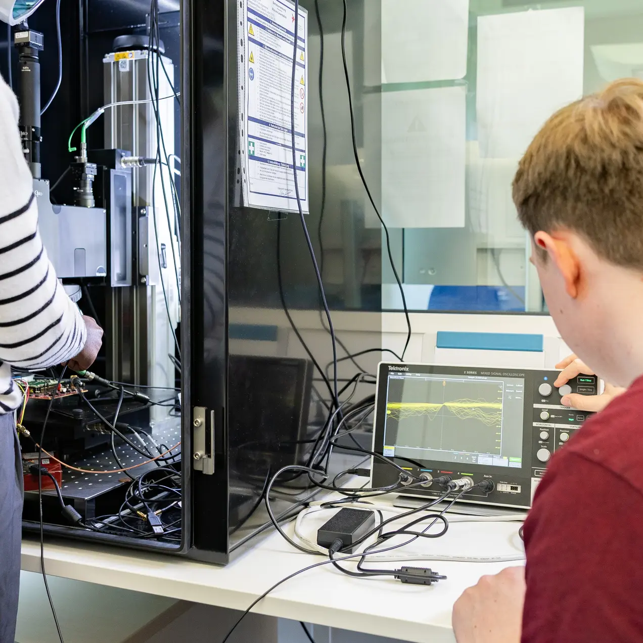 Eine Person mit VR-Brille arbeitet an einem technischen Gerät in einem Schrank, während eine andere Person am Tisch sitzt und Messwerte auf einem Bildschirm überprüft. Beide sind in einem modernen Labor mit Geräten und Kabeln umgeben.