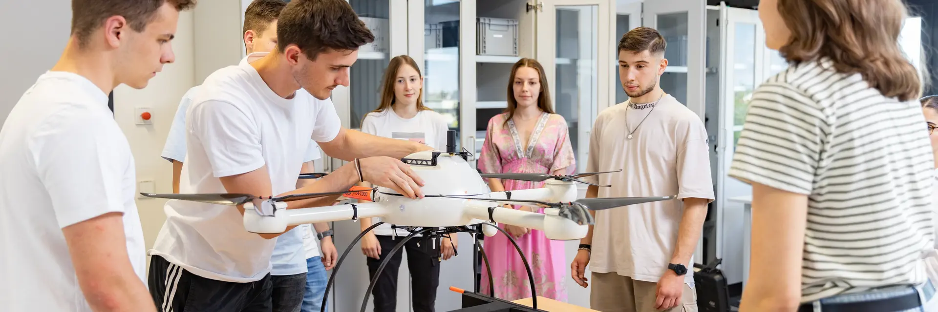 Eine Gruppe von sechs Studierenden steht um einen Tisch, auf dem eine große Drohne platziert ist. Ein Student erklärt anschaulich etwas zur Drohne, während die anderen aufmerksam zuhören und beobachten. Der Raum ist hell und modern eingerichtet.