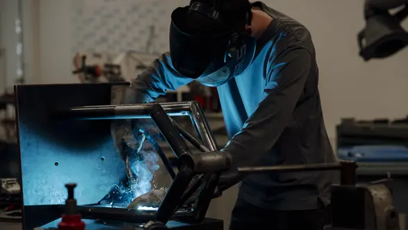 Ein Facharbeiter mit Schweißhelm arbeitet an einem Metallrahmen in einer Werkstatt. Funken und Licht entstehen beim Schweißen, während er präzise an der Metallkonstruktion arbeitet. Werkzeuge und Materialien sind auf dem Tisch verstreut.