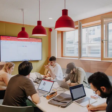 Eine Gruppe von sechs Personen arbeitet in einem hellen Besprechungsraum. Alle haben Laptops geöffnet und sind mit verschiedenen Aufgaben beschäftigt. An einer Wand befindet sich ein Bildschirm, der Informationen anzeigt. Rote Lampen hängen von der Decke.