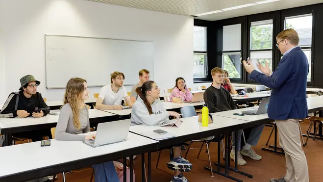 Ein Lehrer steht vorne im Klassenraum und erklärt den Schülern, die an Tischen sitzen und auf Notebooks oder ihre Notizen schauen. Die Schüler wirken interessiert und aufmerksam, während der Lehrer mit Gesten die Erklärung unterstützt. 