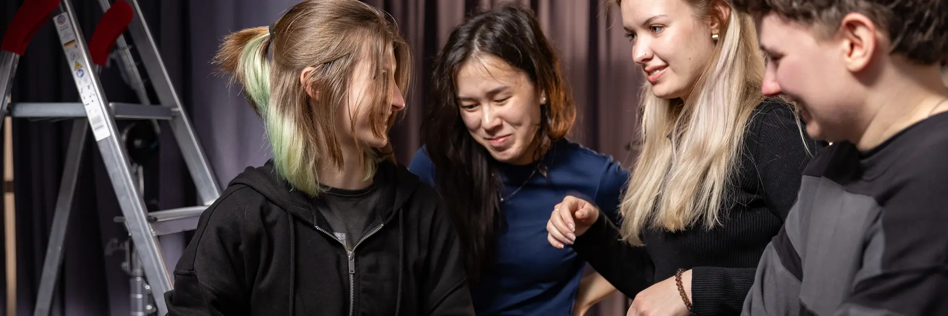 Vier junge Frauen stehen in einem Raum und schauen gemeinsam auf eine Kamera. Eine von ihnen hält die Kamera in der Hand und lächelt, während die anderen interessiert zuschauen. Die Stimmung wirkt freundlich und kooperativ.