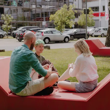 Drei junge Erwachsene sitzen auf einer roten Sitzgelegenheit im Freien. Sie unterhalten sich, lächeln und scheinen eine entspannte Zeit zu verbringen. Im Hintergrund sind einige Autos und ein modernes Gebäude sichtbar.