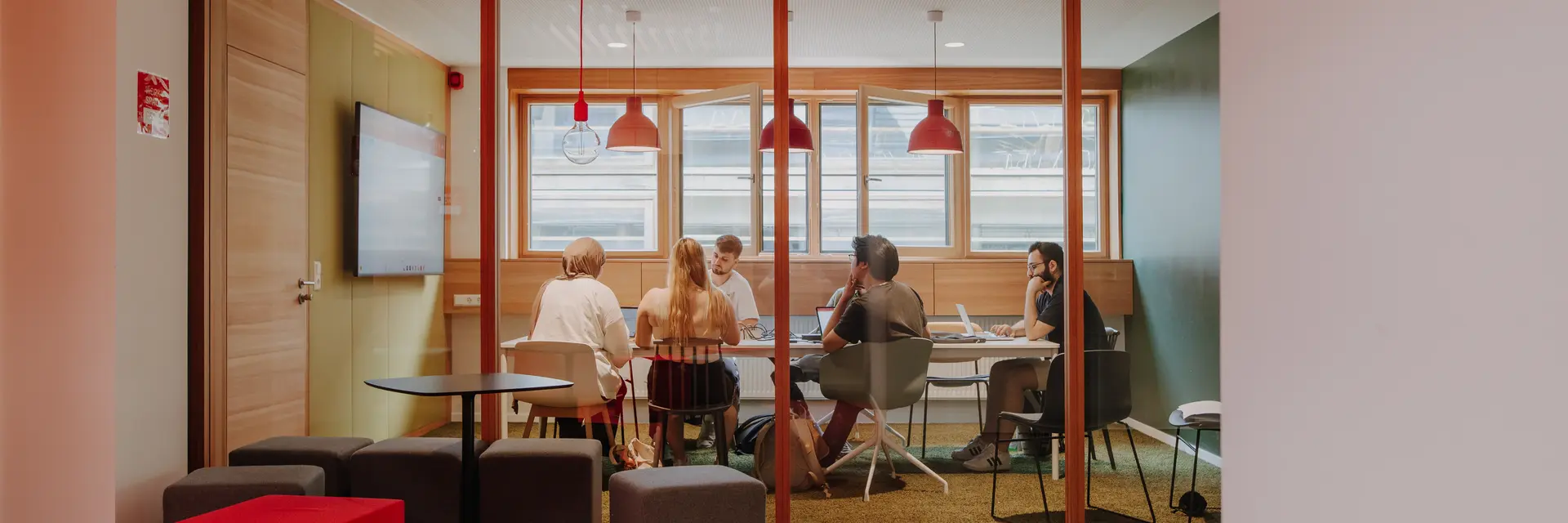 Eine Gruppe von fünf Personen sitzt an einem Tisch in einem modernen Besprechungsraum. Sie sind mit verschiedenen Geräten beschäftigt. Der Raum hat große Fenster und ist hell erleuchtet, während der Boden in einem kräftigen Rot gehalten ist.