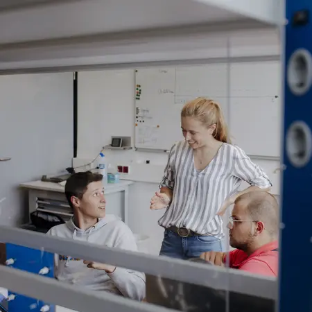 Drei Personen sitzen in einem modernen Büroraum. Eine Frau mit langen, blonden Haaren steht und spricht mit zwei Männern, die an einem Tisch sitzen. Die Atmosphäre wirkt kreativ und kommunikativ.