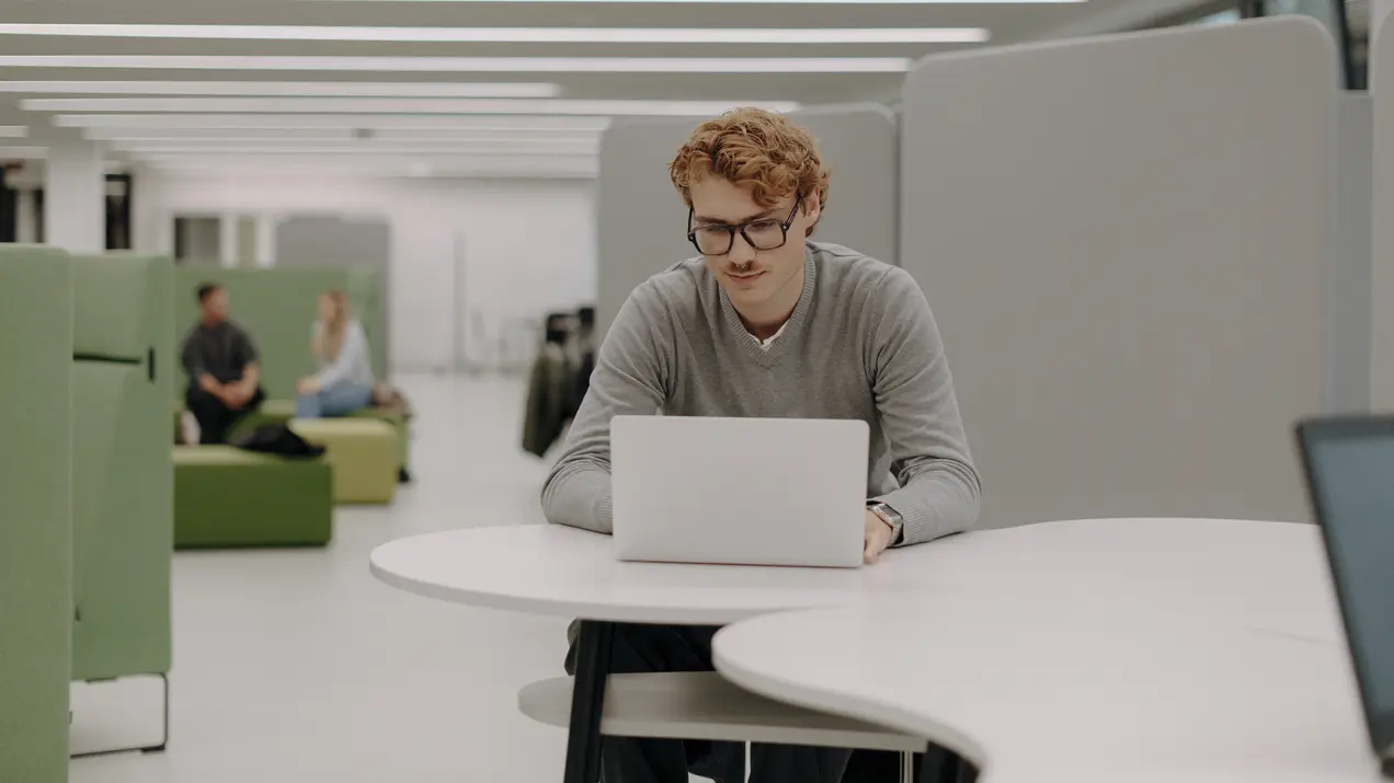 Ein Mann mit Locken und Brille sitzt an einem Tisch und arbeitet konzentriert an einem Laptop. Im Hintergrund sind weitere Personen zu sehen, die in einer modernen, hellen Umgebung miteinander sprechen oder entspannen.