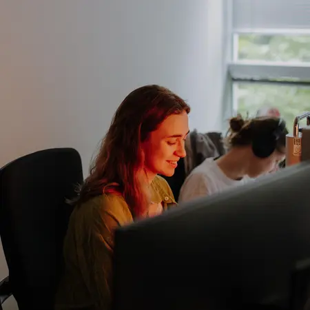 Eine Frau sitzt an einem Schreibtisch vor einem Computerbildschirm. Sie schaut konzentriert auf den Bildschirm. Im Hintergrund ist eine weitere Person zu sehen, die Kopfhörer trägt. Das Licht im Raum hat einen warmen Farbton.