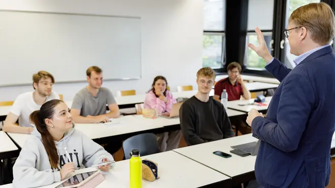 Eine Lehrperson steht vor einer Gruppe von Studierenden in einem Klassenzimmer. Die Studierenden sitzen an Tischen, einige schauen aufmerksam, andere machen Notizen. Die Atmosphäre ist konzentriert und lehrreich.