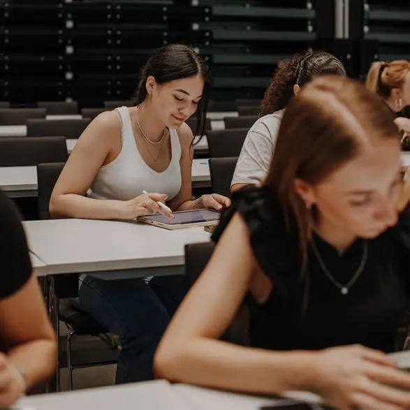 Eine junge Frau in einem weißen Tanktop sitzt an einem Tisch und schreibt in ein Notizbuch. Im Hintergrund sind weitere Studierende zu sehen, die konzentriert an ihren Aufgaben arbeiten. Der Raum hat eine moderne, minimalistische Gestaltung.