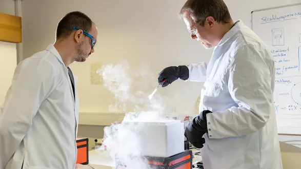 Zwei Wissenschaftler in Laborkittel stehen in einem Labor. Einer der Wissenschaftler arbeitet mit einer mit Nebel gefüllten Box und hält einen Löffel. Der andere Wissenschaftler beobachtet den Vorgang. Beide tragen Schutzbrillen und Handschuhe.