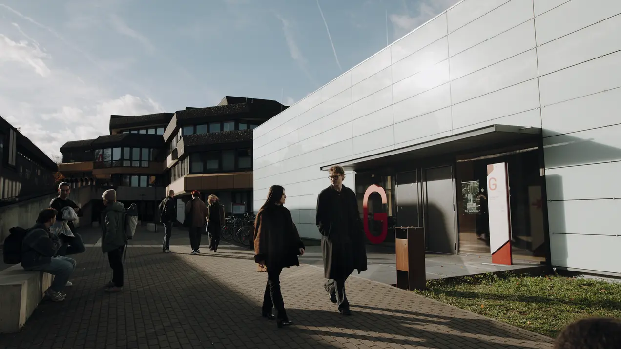 Eine Gruppe von Menschen geht auf einem Platz vor modernen Gebäuden. Im Vordergrund sind zwei Personen im Gespräch. Das Licht ist sanft, und der Himmel ist teilweise bewölkt. Das Ambiente wirkt lebhaft und einladend.