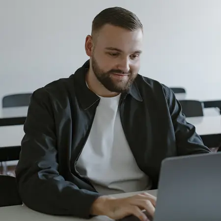 Ein Mann mit Bart sitzt an einem Tisch und arbeitet auf einem Laptop. Er trägt eine dunkle Jacke und ein weißes T-Shirt. Seine Miene ist konzentriert und er tippt auf der Tastatur, während er auf den Bildschirm schaut.