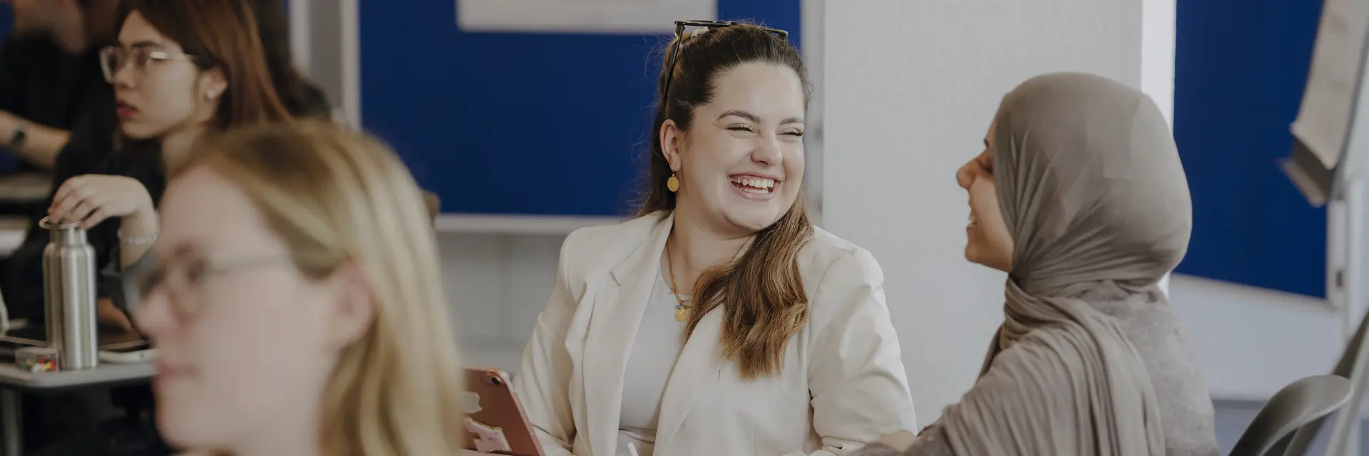 Zwei Frauen sitzen an einem Tisch und lachen miteinander. Eine Frau trägt einen hellen Blazer, während die andere ein Kopftuch trägt. Im Hintergrund sind verschwommen andere Personen und teilweise weiße Wände mit blauen Pinnwänden zu sehen.