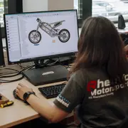 EinePerson sitzt vor einem Computer und arbeitet an einem digitalen Modell eines Motorradrahmens. Der Bildschirm zeigt die 3D-Ansicht des Rahmens, während die Person auf die Elemente konzentriert ist. Der Arbeitsbereich ist gut beleuchtet und modern gestaltet.