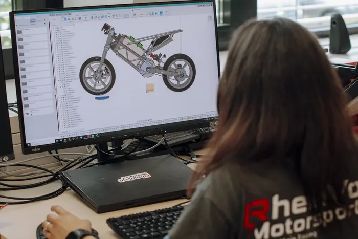 EinePerson sitzt vor einem Computer und arbeitet an einem digitalen Modell eines Motorradrahmens. Der Bildschirm zeigt die 3D-Ansicht des Rahmens, während die Person auf die Elemente konzentriert ist. Der Arbeitsbereich ist gut beleuchtet und modern gestaltet.