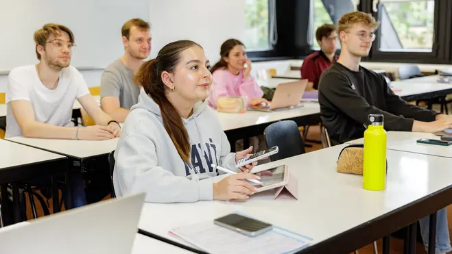 Eine Gruppe von sechs jungen Menschen sitzt in einem Klassenzimmer. Einige nutzen Laptops, andere halten Smartphones oder Notizen in der Hand. Die Atmosphäre wirkt konzentriert und lernend, während die Schüler aufmerksam nach vorne schauen.