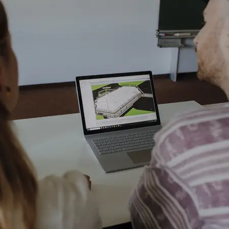Zwei Personen sitzen an einem Tisch und betrachten einen Laptop. Auf dem Bildschirm ist ein 3D-Modell eines Gebäudes zu sehen. Die Personen scheinen interessiert und diskutieren über das gezeigte Modell.