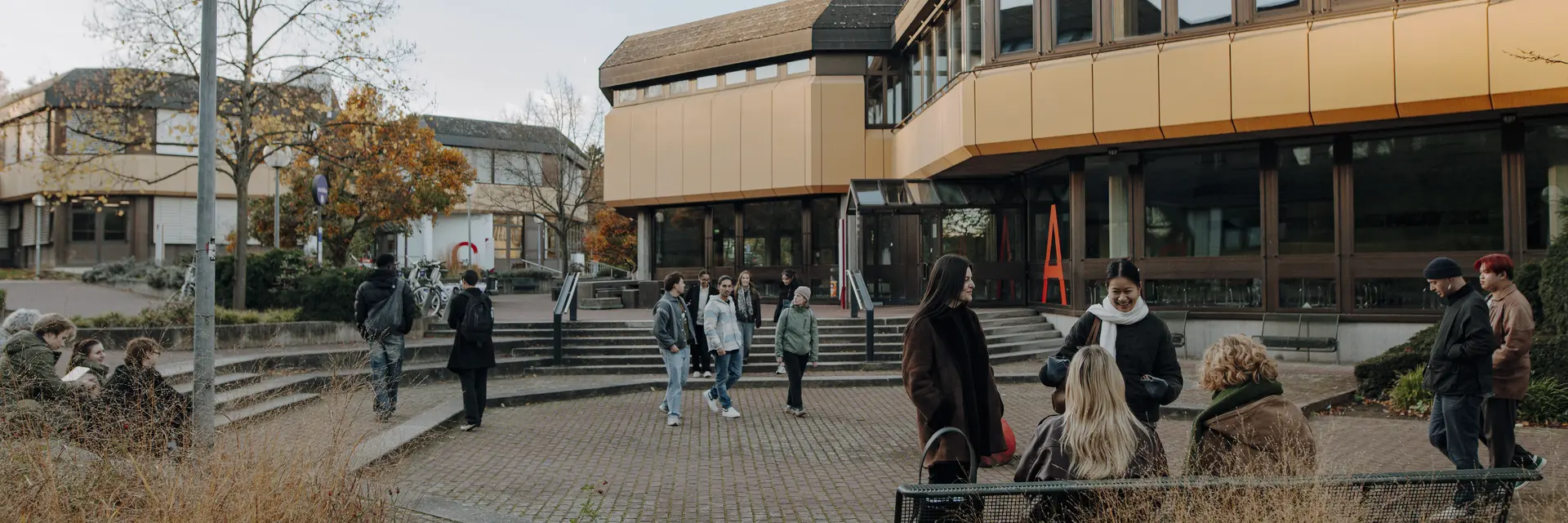 Eine Gruppe von Personen steht und sitzt vor einem modernen, zweigeschossigen Gebäude mit großen Fenstern. Einige Menschen unterhalten sich, während andere auf einer Bank sitzen. Umgebende Pflanzen und Bäume verleihen der Szenerie eine natürliche Atmosphäre.