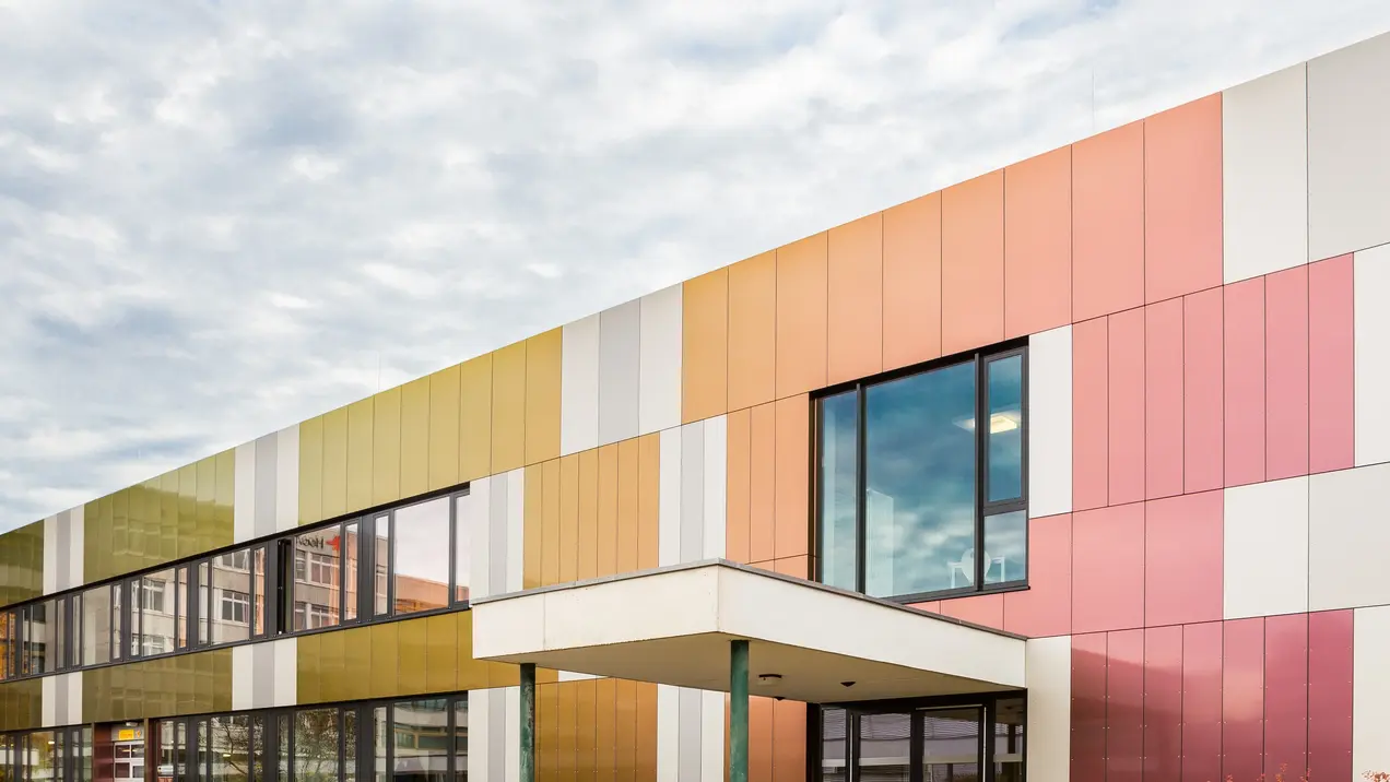 Ein modernes Gebäude mit einer farbenfrohen Fassade, die aus verschiedenen Paneelen in Gelb-, Rosa- und Weißtönen besteht. Die Fenster sind groß und bieten einen Blick auf die Umgebung. Der Himmel ist bewölkt, was dem Bild eine ruhige Atmosphäre verleiht.
