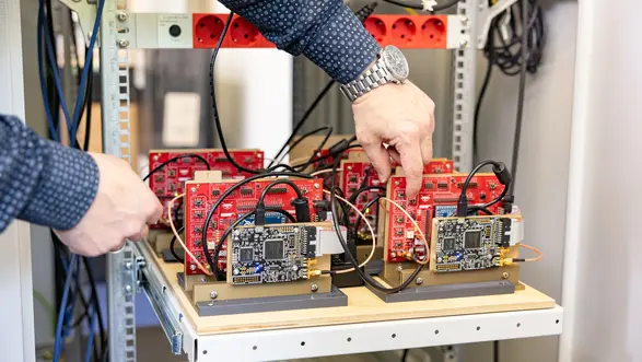 Eine Person in einem blauen Hemd arbeitet an einem Serverrack. Sie berührt eine Platine, während mehrere rote Platinen und Kabel im Rack sichtbar sind. Das Bild zeigt eine technische Umgebung, in der elektronische Komponenten installiert oder gewartet werden.