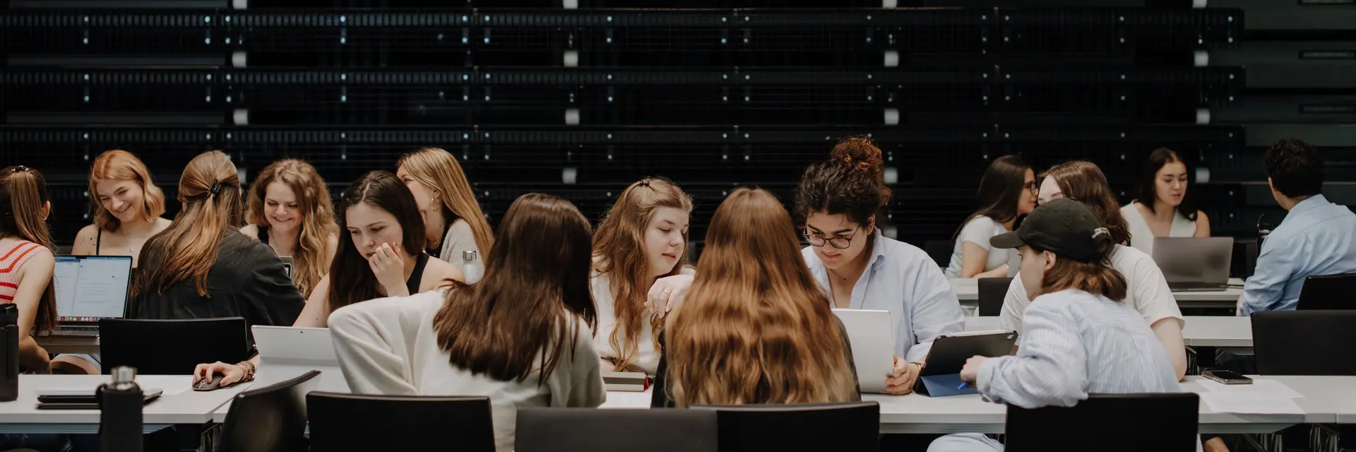 Eine Gruppe von Studierenden, hauptsächlich Frauen, sitzt an Tischen in einem Raum mit dunklen Wänden. Sie arbeiten konzentriert an Laptops und führen Gespräche. Die Atmosphäre wirkt lebhaft und kooperativ.