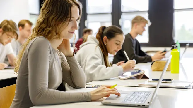Eine Gruppe von Studierenden sitzt in einem Klassenzimmer. Eine Studentin konzentriert sich auf ihren Laptop, während eine andere etwas auf einem Tablet schreibt. Im Hintergrund sind weitere Studierende zu sehen, die ebenfalls am Arbeiten sind.