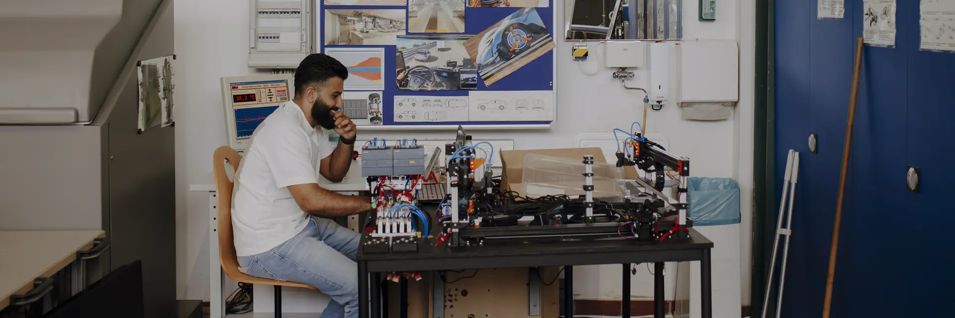Ein Mann sitzt an einem Tisch in einem technischen Raum. Vor ihm befindet sich ein elektronisches Gerät und er hält nachdenklich einen Kaffeebecher in der Hand. Im Hintergrund hängt eine Wandtafel mit technischen Zeichnungen und Plänen.
