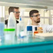 Zwei Wissenschaftler in Schutzbrillen arbeiten in einem Labor. Einer von ihnen hält ein Glasgefäß, während der andere aufmerksam zusieht. Im Hintergrund sind verschiedene Laborutensilien und Chemikalien sichtbar.