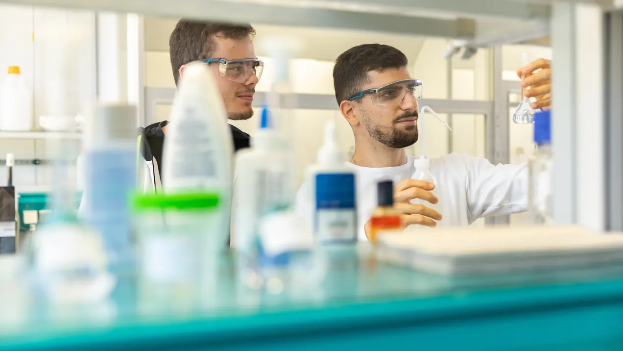 Zwei Wissenschaftler in Schutzbrillen arbeiten in einem Labor. Einer von ihnen hält ein Glasgefäß, während der andere aufmerksam zusieht. Im Hintergrund sind verschiedene Laborutensilien und Chemikalien sichtbar.
