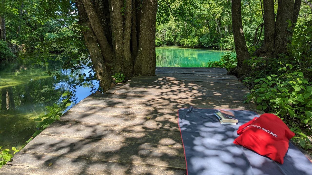 Es ist ein Holzsteg zu sehen, auf dem ein Handtuch mit Buch und HSRM-Tasche liegt. Ein türkiser Fluss im Hintergrund schimmert in der Sonne
