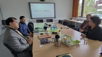 Ein Tisch mit fünf Personen in einem Konferenzraum. Einige Teilnehmer diskutieren, während ein Bildschirm mit der Aufschrift „Marokko - Deutschland“ im Hintergrund sichtbar ist. Auf dem Tisch stehen Getränke und Unterlagen, die zur Besprechung genutzt werden.