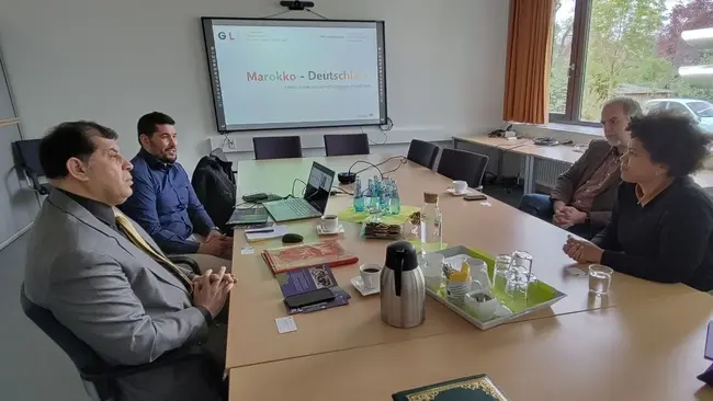 Ein Tisch mit fünf Personen in einem Konferenzraum. Einige Teilnehmer diskutieren, während ein Bildschirm mit der Aufschrift „Marokko - Deutschland“ im Hintergrund sichtbar ist. Auf dem Tisch stehen Getränke und Unterlagen, die zur Besprechung genutzt werden.