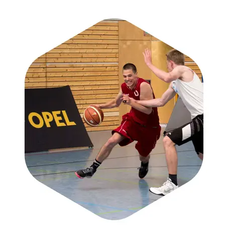Ein Basketballspieler in rotem Trikot dribbelt den Ball und versucht, einem verteidigenden Spieler in einem weißen Trikot zu entkommen. Die Szene spielt sich in einer Halle mit Holzvertäfelung ab.