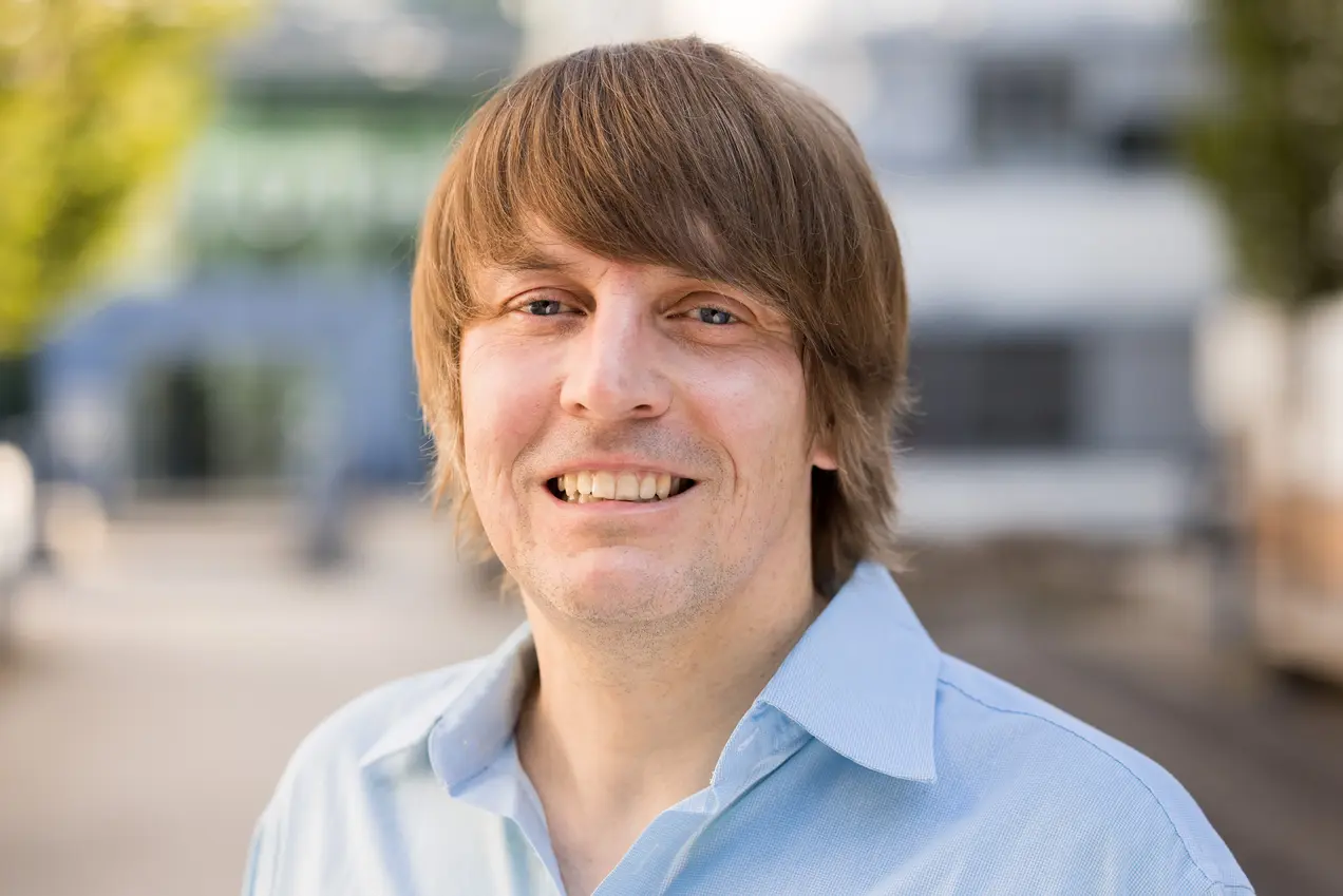 Ein lächelnder Mann mit langen, blonden Haaren trägt ein hellblaues Hemd. Er steht im Freien, mit verschwommenem Hintergrund von Gebäuden. Das Bild vermittelt eine freundliche und einladende Stimmung.