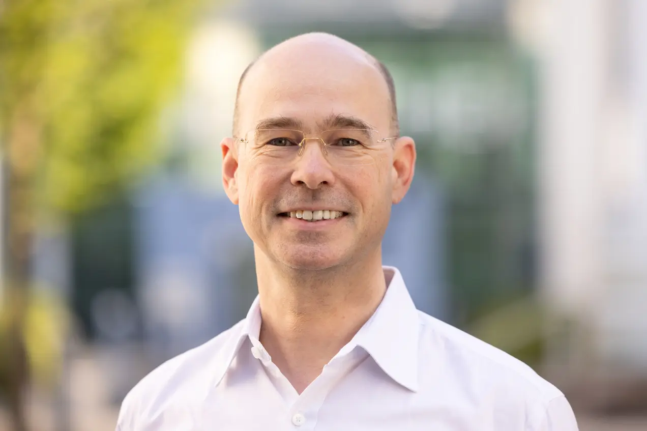 Ein lächelnder Mann mit Glatze trägt eine Brille und ein weißes Hemd. Er steht draußen vor einer unscharfen Hintergrundlandschaft mit grünen und hellen Elementen. Der Ausdruck ist freundlich und einladend.