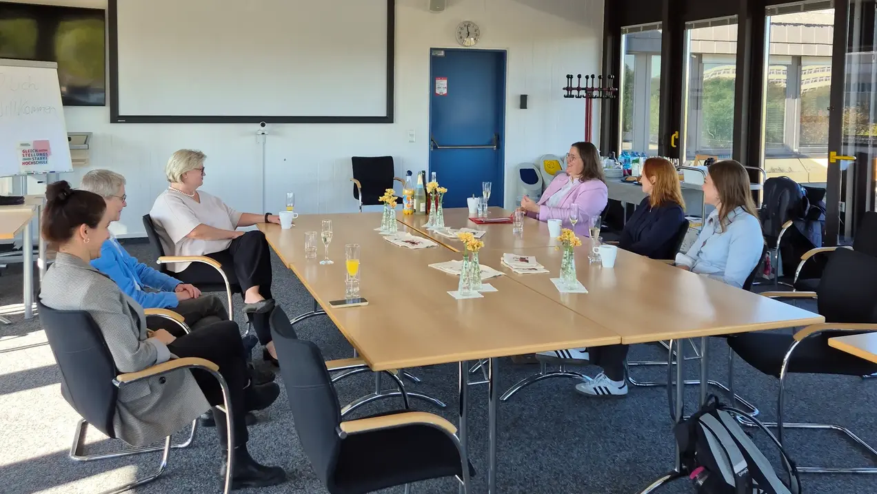 Eine Gruppe von sieben Personen sitzt an einem langen Tisch in einem Konferenzraum. Auf dem Tisch stehen Vasen mit Blumen und einige Gläser. Eine Frau spricht, während die anderen aufmerksam zuhören. Im Hintergrund sind Fenster zu sehen, die Tageslicht in den Raum lassen.