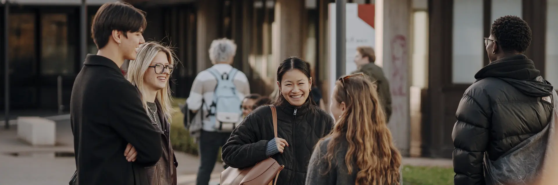 Sechs junge Menschen stehen in einem Freiraum, lächeln und unterhalten sich. Einige tragen modische Kleidung und das Umfeld wirkt urban und freundlich. Im Hintergrund sind weitere Personen und Gebäude zu erkennen, was auf eine lebendige Atmosphäre hindeutet.