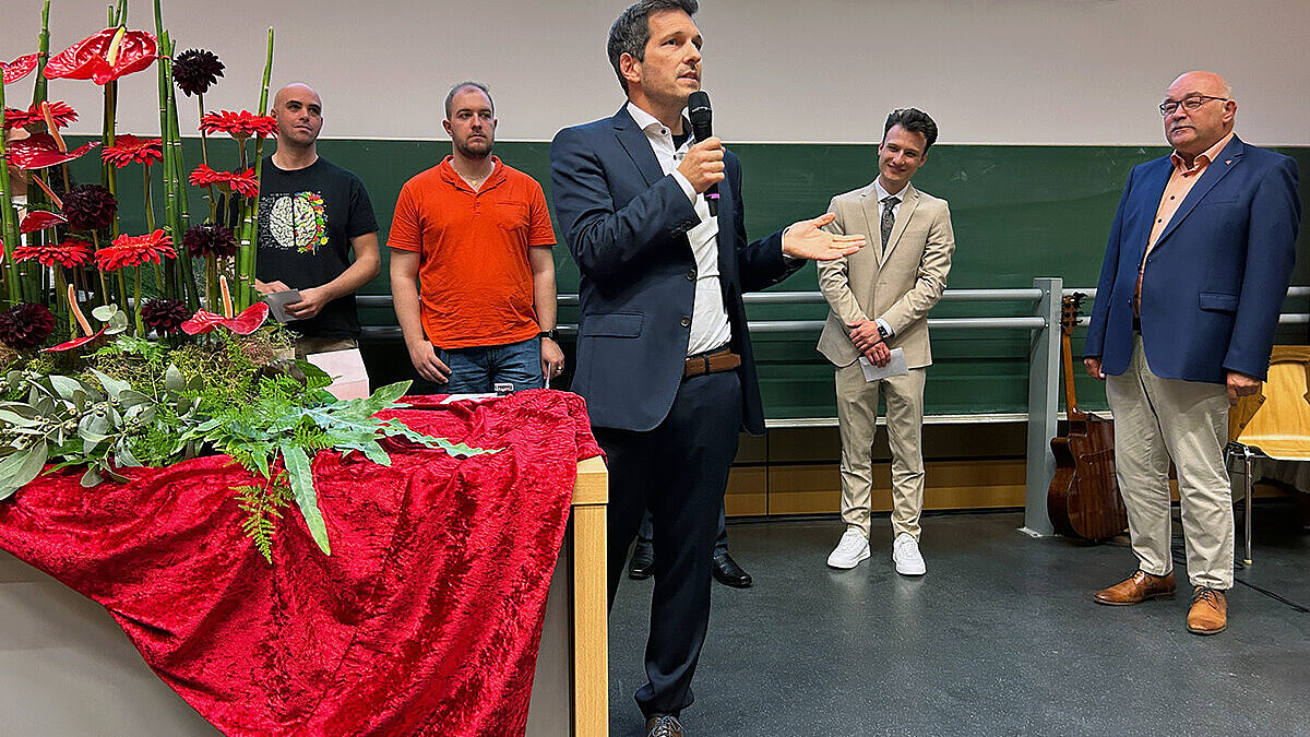 Ein Mann mit Mikrophon spricht zum Publikum. Dahinter stehen weitere Personen.