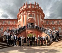 Auf einer großen Freutreppe eines Schlosses stehen Menschen und posieren für ein Gruppenfoto.