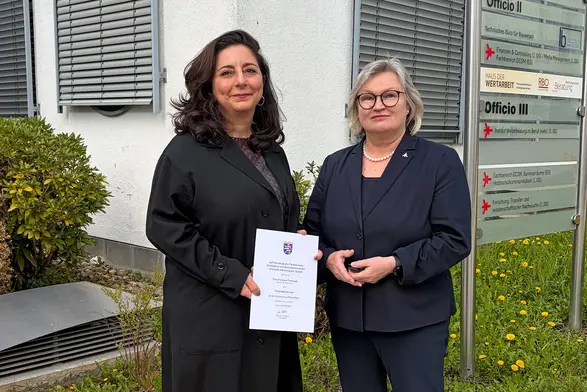 Zwei Frauen stehen im Freien vor einem Gebäude, jede hält ein Dokument in der Hand. Die Frau links trägt einen schwarzen Mantel und hat lockige Haare, während die Frau rechts einen dunklen Blazer trägt und eine Brille. Im Hintergrund sind Fenster und eine Pflanze sichtbar.