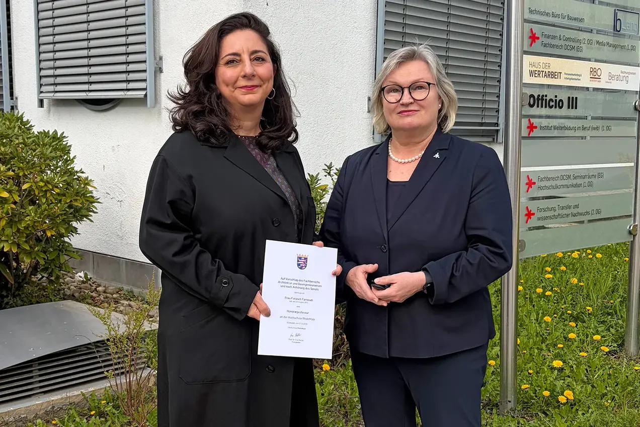 Zwei Frauen stehen im Freien vor einem Gebäude, jede hält ein Dokument in der Hand. Die Frau links trägt einen schwarzen Mantel und hat lockige Haare, während die Frau rechts einen dunklen Blazer trägt und eine Brille. Im Hintergrund sind Fenster und eine Pflanze sichtbar.