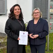 Zwei Frauen stehen im Freien vor einem Gebäude, jede hält ein Dokument in der Hand. Die Frau links trägt einen schwarzen Mantel und hat lockige Haare, während die Frau rechts einen dunklen Blazer trägt und eine Brille. Im Hintergrund sind Fenster und eine Pflanze sichtbar.