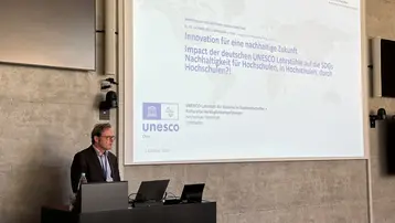 Prof. Dr.-Ing. Michael Kloos steht an einem Podium und präsentiert auf einer großen Leinwand seine Arbeit am UNESCO Lehrstuhl der HSRM. Der Inhalt behandelt außerdem die Rolle von Hochschulen in der Umsetzung von nachhaltigen Entwicklungszielen.